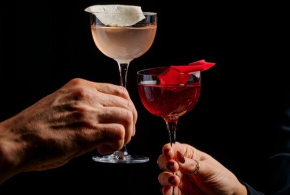 Hands clinking two cocktails, one pale, the other red.