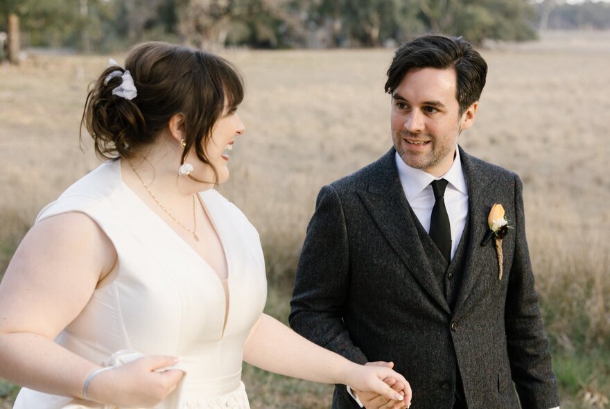 A wedding couple holding hands in a field, looking at each other and smiling.