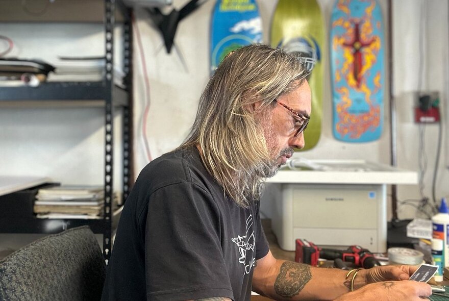 An artist seated at a desk, working on a collage, with three colourful skateboards displayed on the wall behind him, next to black shelves.