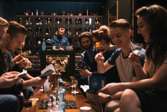 A group of people seated around a low table taking part in an interactive cocktail masterclass, a bartender in the background.