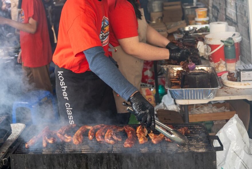 Cook holding tongs and wearing gloves turning skewers on a smoking-hot griddle.