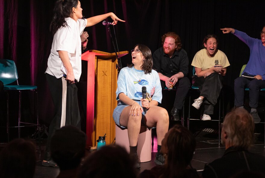 Six comedians on stage all laughing, one seated on a pink stool.
