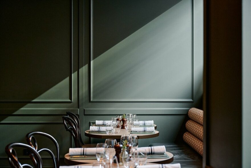 View of empty tables and chairs in a restaurant, ready for service with plates, napkins, glassware and cutlery, and soft light from a window shining on a pale green wall.