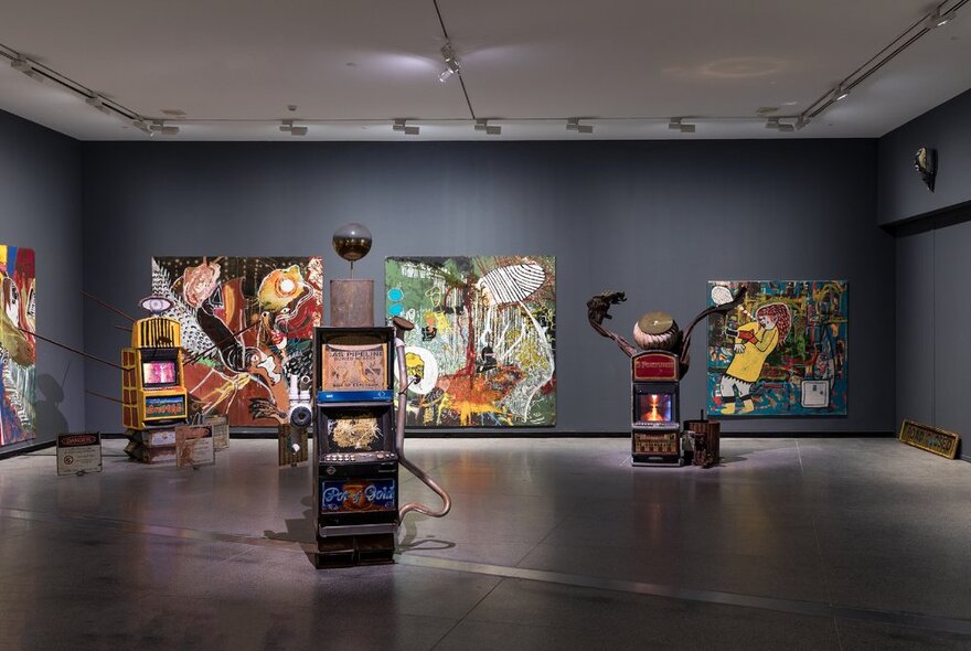 An exhibition space at a gallery with Indigenous paintings displayed on the walls and sculptures placed on the concrete floor.