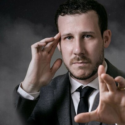 Magician and mentalist, Tom Weil, wearing a suit and with a serious expression, one hand resting on his temple, the other hand stretched out towards the viewer.