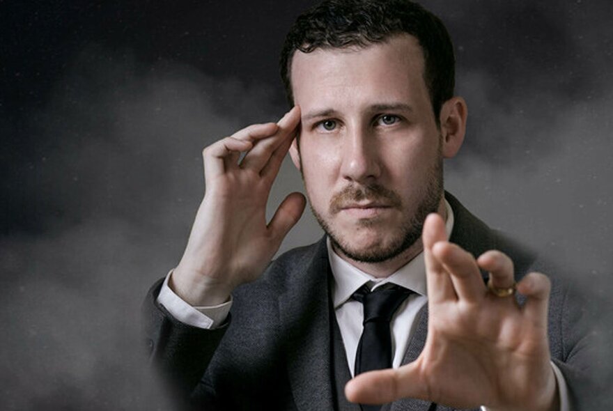 Magician and mentalist, Tom Weil, wearing a suit and with a serious expression, one hand resting on his temple, the other hand stretched out towards the viewer.