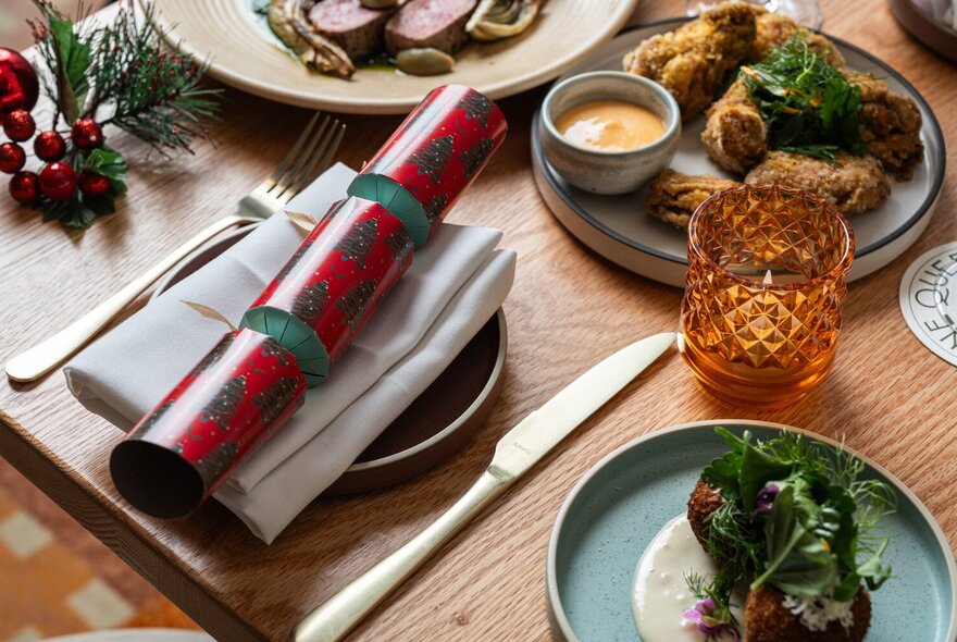 A place setting at a table, with a napkin and Christmas cracker resting on the plate, with plates of food also on the table.