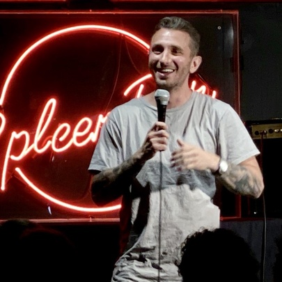 Stand-up comedian Tommy Little on stage in front of Spleen neon sign, holding microphone and smiling.
