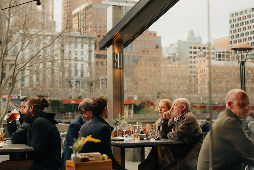 Patrons seated outside at a restaurant on the banks of the Yarra River in winter, the buildings on the opposite bank behind them.