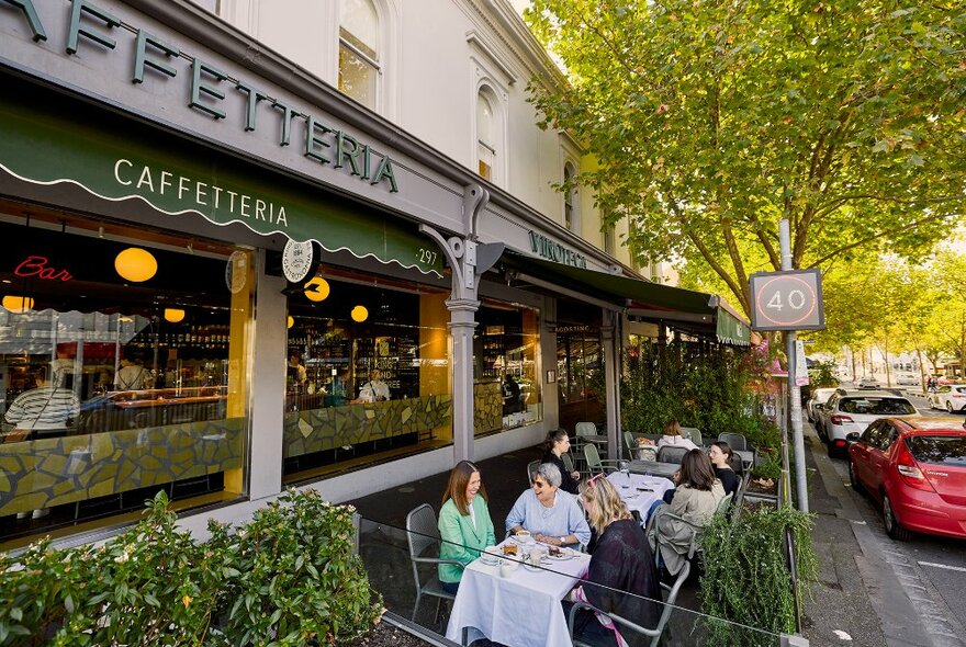 A group of women sitting at a table outside a caffetteria surrounded by green trees and plants.