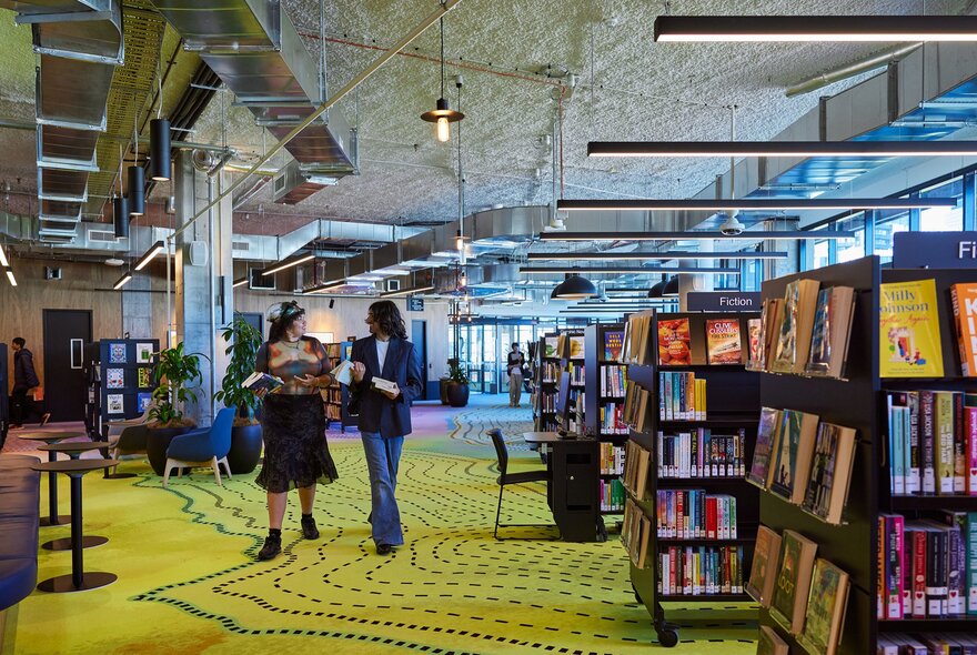 Two friends are walking through a library. There is colourful artwork on the carpet and shelves of books.