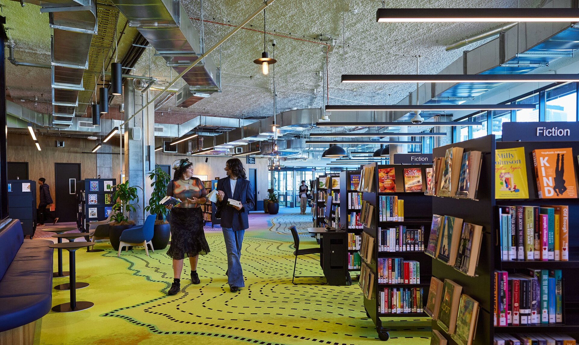 Two friends are walking through a library. There is colourful artwork on the carpet and shelves of books.