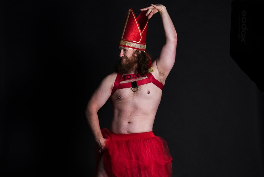 A shirtless man in a red cardinal's hat and red skirt poses with one hand raised above his head. 