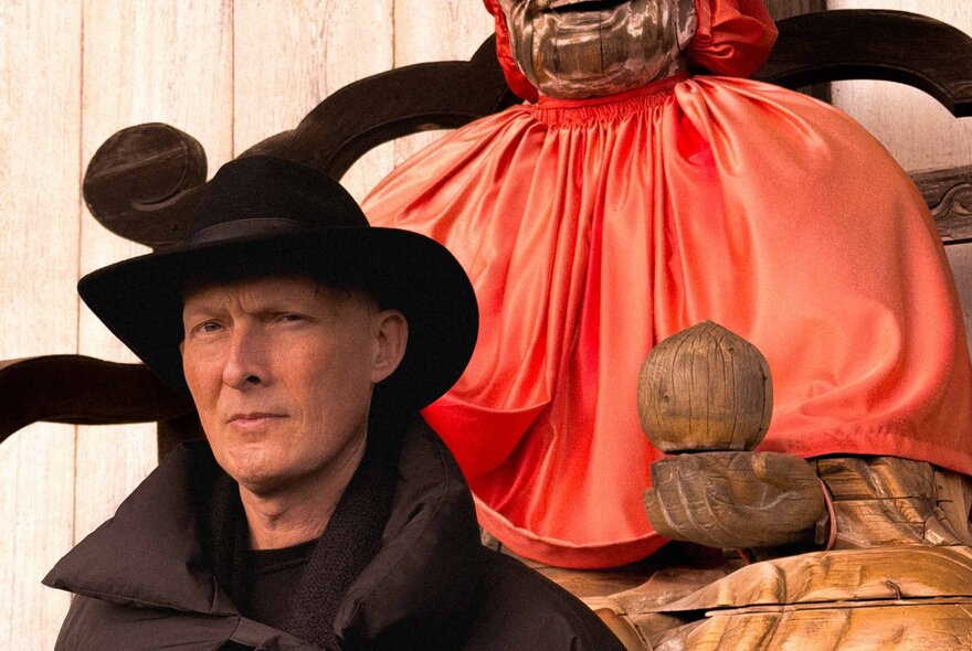 A man in a large had looking stern in front of a sculpture with orange/pink material draped over it. 