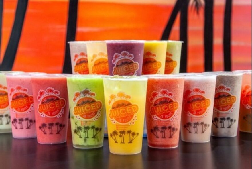 A double row of colourful juices in plastic takeaway cups, all branded with the Juiced logo, arranged on a table.