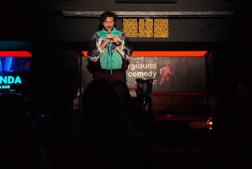 A person on a stage, performing to a seated live audience at a venue hosting stand-up comedy.