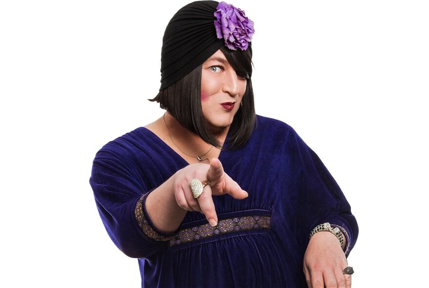 Cabaret artist Tina Del Twist, wearing a purple gown and flower in their hair, pointing a finger and looking coy.