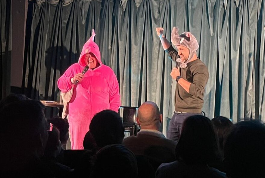 Two performers on a small stage in front of a live audience, dressed in animal costumes and talking into microphones.