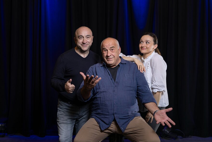 Three prominent figures from the Australian Greek comedy scene posing on a theatre stage in casual clothes.