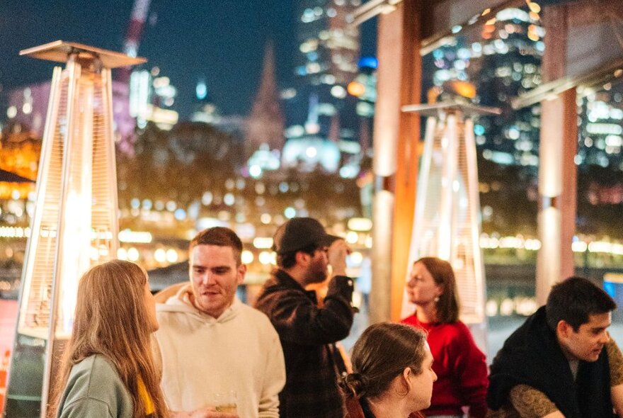 People at a bar with lit up terrace heaters behind them and Melbourne's city lights in the background.
