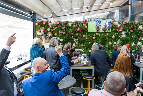 The back of people in formal clothing watching the Melbourne Cup on a tv screen, roses on the back wall. 