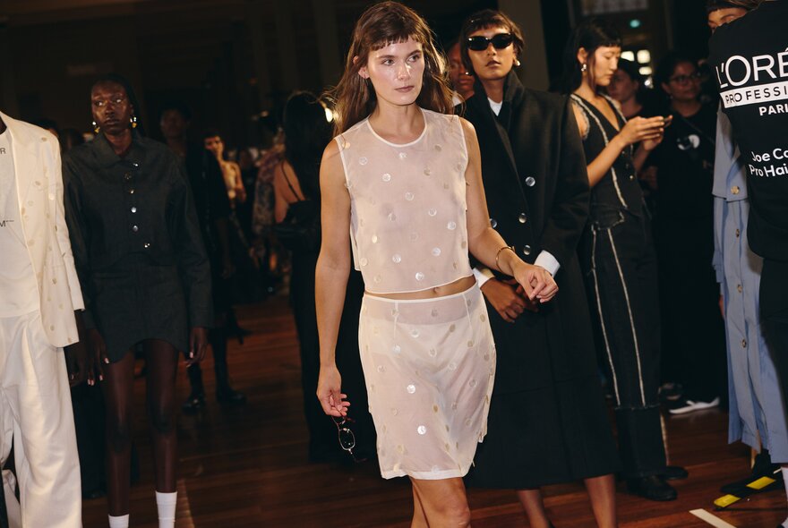 A model walks down a fashion runway wearing a sheer, sleeveless pink top and a matching pink skirt, with blurred figures in the background.