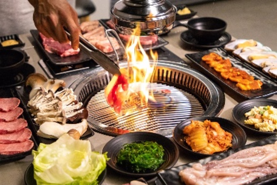 Person using tongs to hold a piece of meat over an open flame at the centre of a small grill BBQ on a dining table at a Korean BBQ restaurant, with plates of fresh raw food displayed around the grill plate.