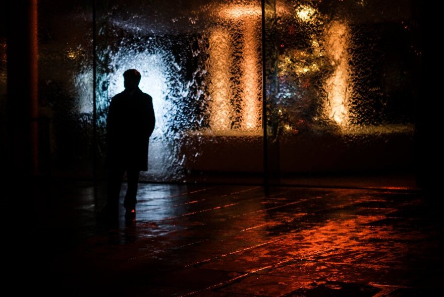 Moody night-time short of a person standing in front of windows in the rain.