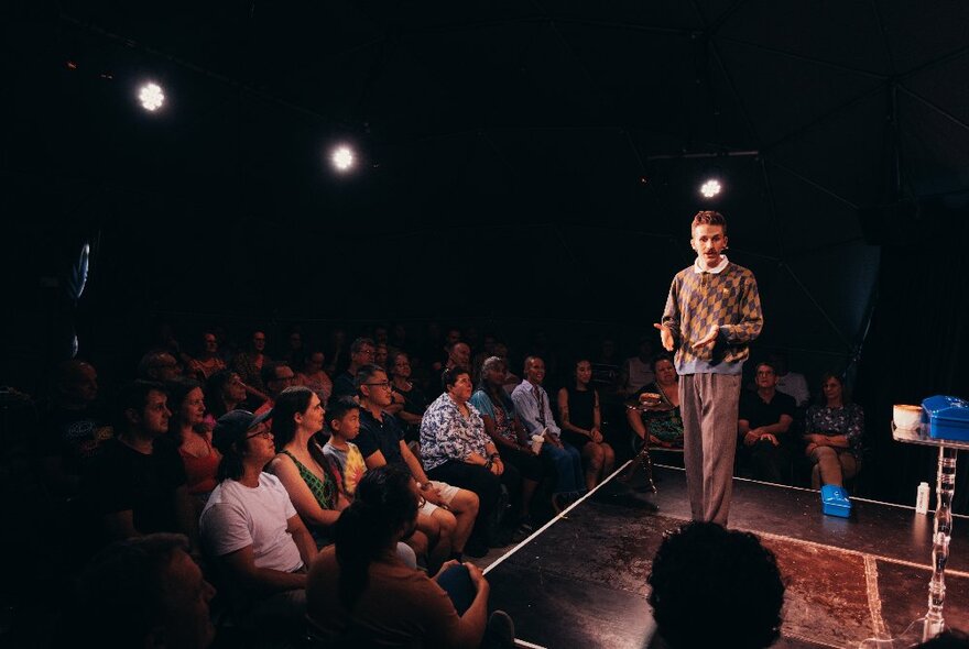 A man on stage wearing a checked jumper, talking to an audience.