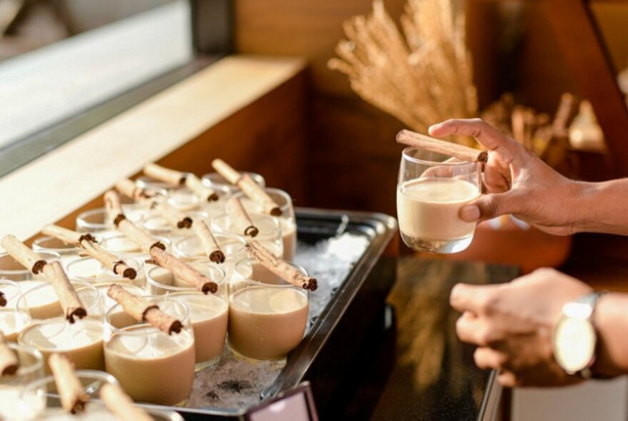 A pair of hands placing a glass of chai (milky tea) onto a tray filled with many glasses of chai, with cinnamon sticks resting on the top of the glasses.