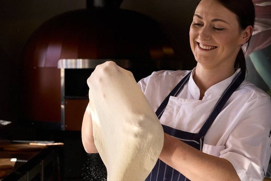 A chef wearing a white chef's shirt and blue striped apron stretching a batch of pizza dough.