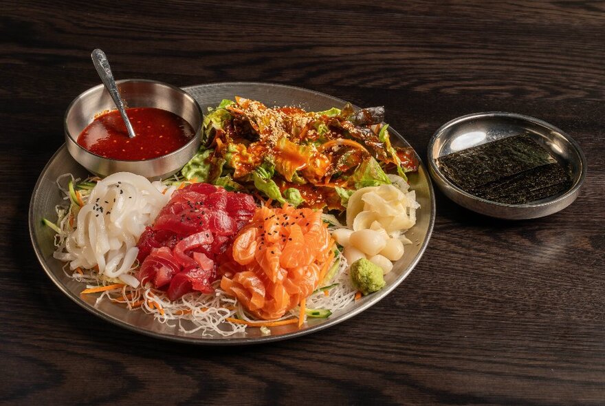 A silver tray containing pieces of raw salmon, tuna and white fish, with a spicy sauce and strips of seaweed.