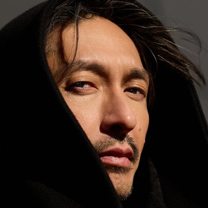 Close-up portrait of the musician Bigyuki (Masayuki Hirano) looking toward the camera while wearing a dark, oversized black hood that frames his face and partially covers his head.