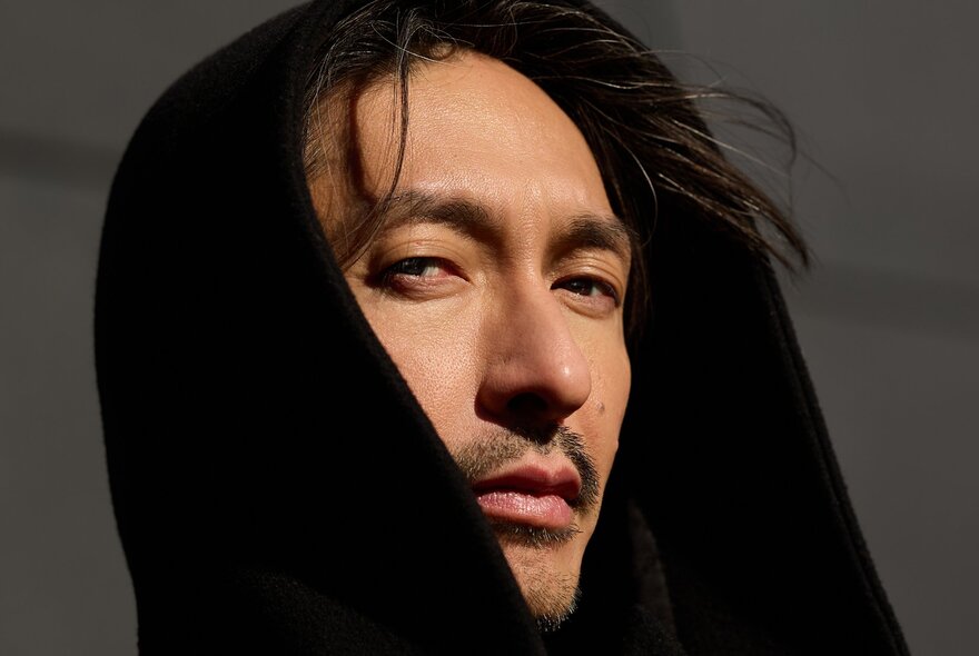 Close-up portrait of the musician Bigyuki (Masayuki Hirano) looking toward the camera while wearing a dark, oversized black hood that frames his face and partially covers his head.