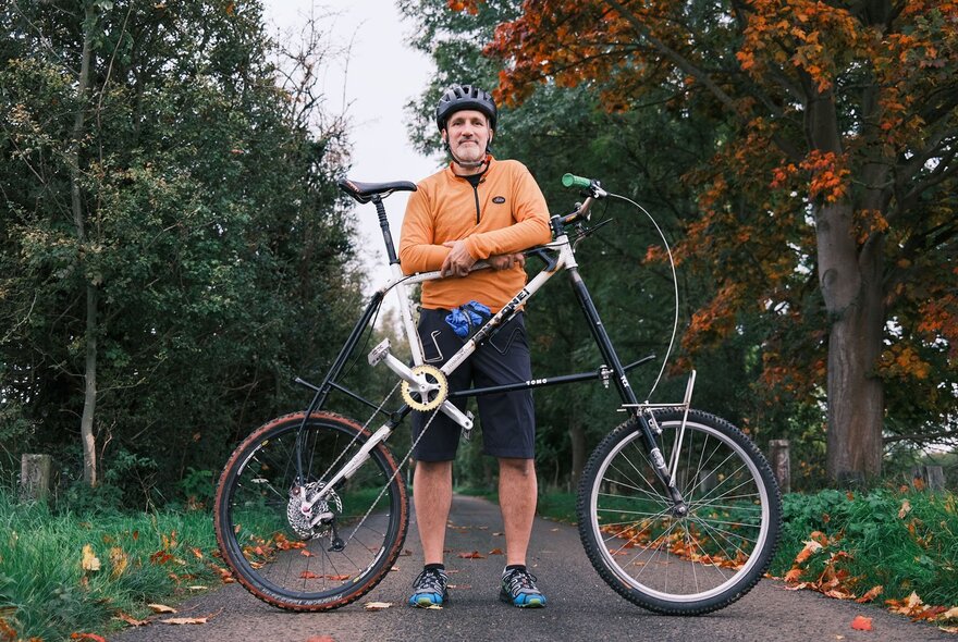 A middle-aged man wearing an orange top, black shorts and a bike helmet, standing behind his tall bike on a path in an outdoor setting. 