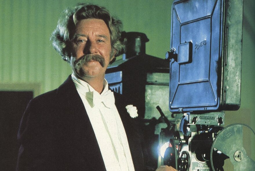 Actor John Meillon wearing tuxedo standing next to old-fashioned cinema equipment.