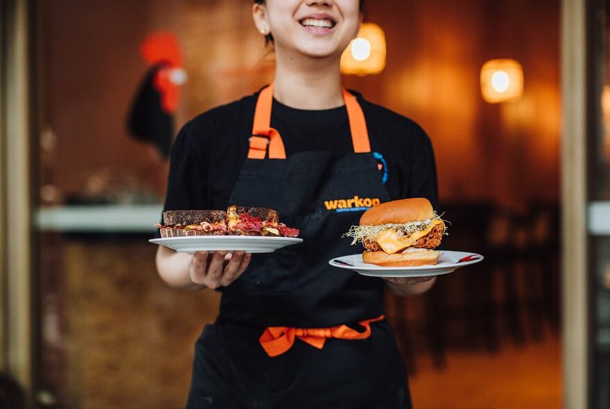 A waiter in an apron holds two plates containing large sandwiches.