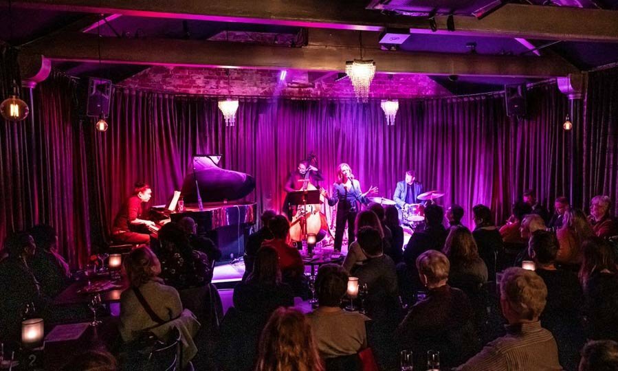 Audience seated in a darkened room watching a band perform on a purple-lit stage. 