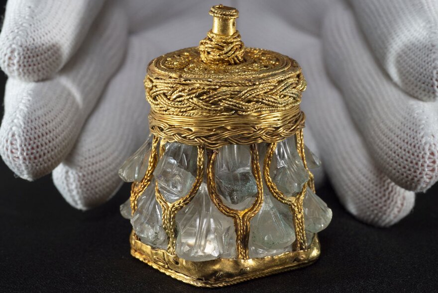 White gloved hands surround a tiny gold and glass piece of Viking treasure. 