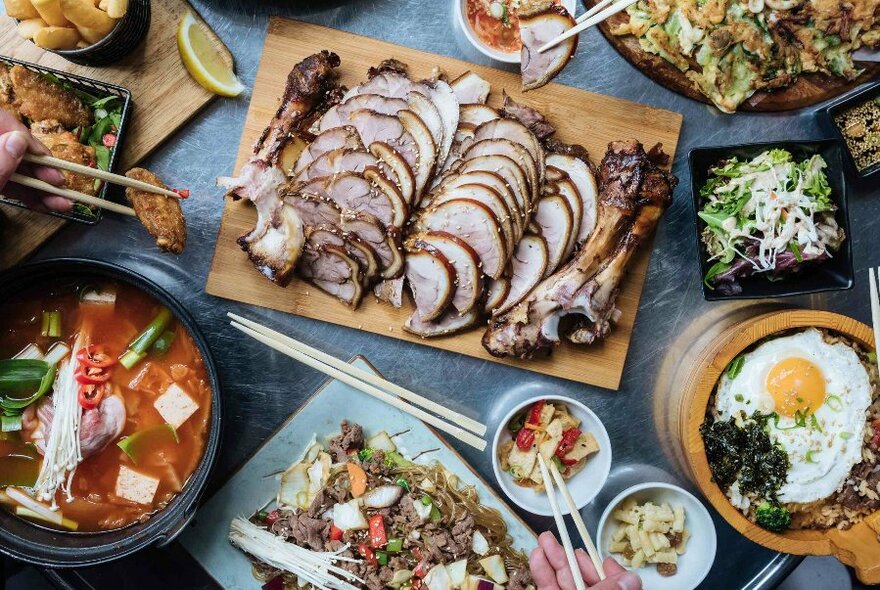 Overhead view of a table filled with plates of Korean food and hands with using chopsticks to select food.