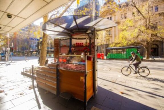 Creperie cart in winter sunshine with dappled light through trees as a cyclist passes on an empty street.