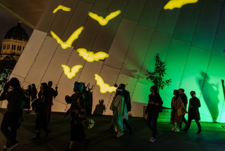 People walking past the walls of the Melbourne Museum with projections of bats in yellow. 