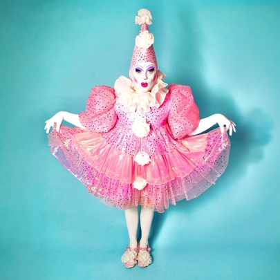 Full-length studio portrait of a performer dressed as a stylised, theatrical pink clown against a solid light blue background. 
