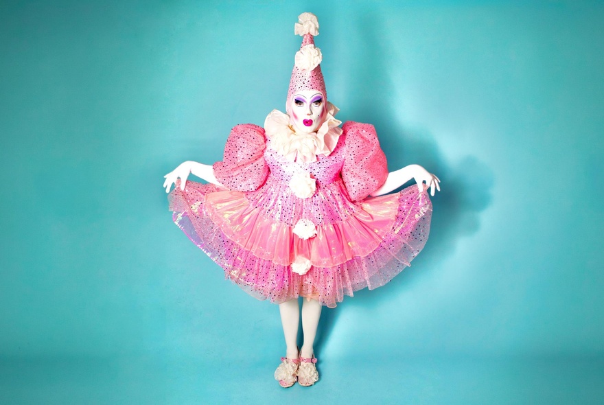 Full-length studio portrait of a performer dressed as a stylised, theatrical pink clown against a solid light blue background. 