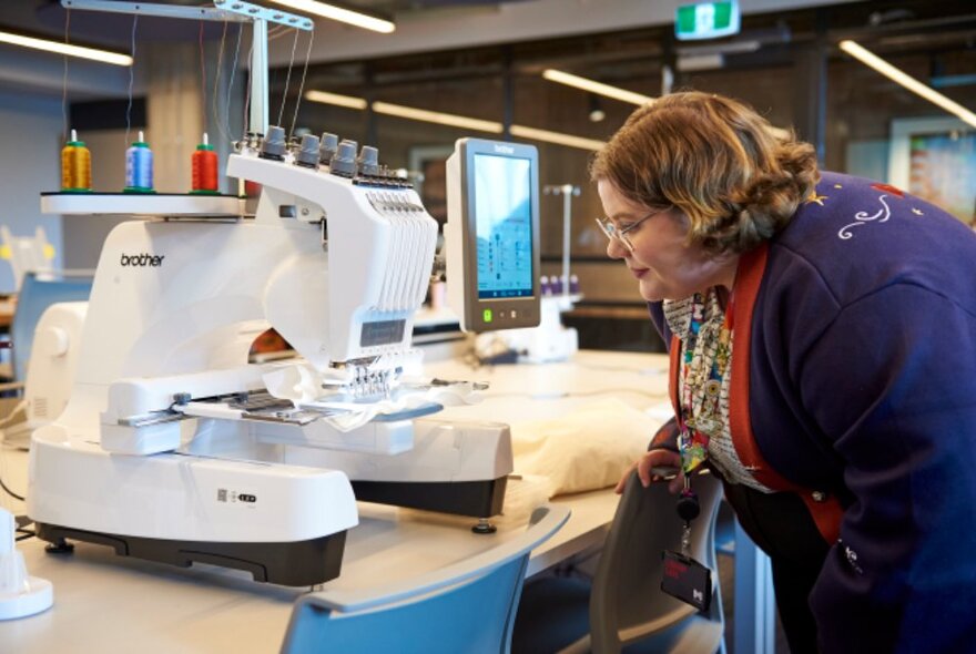 A person looking intently at a sewing or monogramming machine with multiple spools of coloured thread attached to it, on a long table in a room.