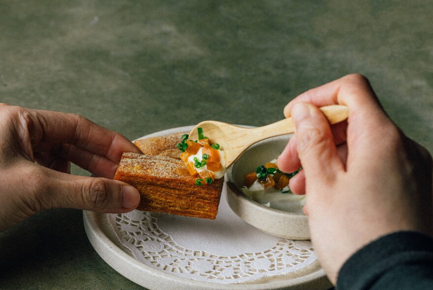 Hand holding a wooden spoon to scoop topping onto a slice over a plate with lace doily.