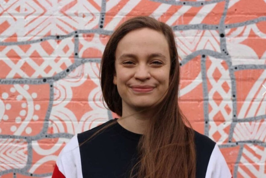 A woman smiles warmly in front of a painted brick wall with patterns. 