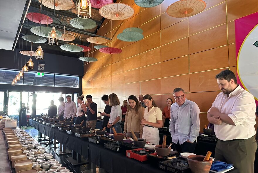 People lined up at workstations with Asian cooking equipment in a restaurant with ceiling hung with paper umbrellas.