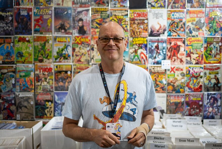 A man wearing a expo lanyard around his neck, standing in front of a booth of comic memorabilia at The Games Expo.