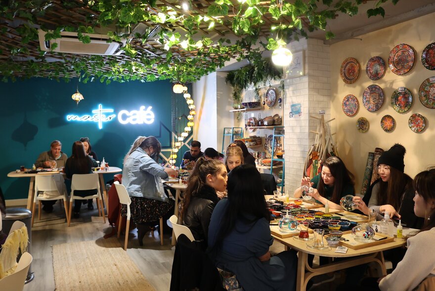 A craft workshop with people at tables working on projects, he roof is made of fake green vines. 